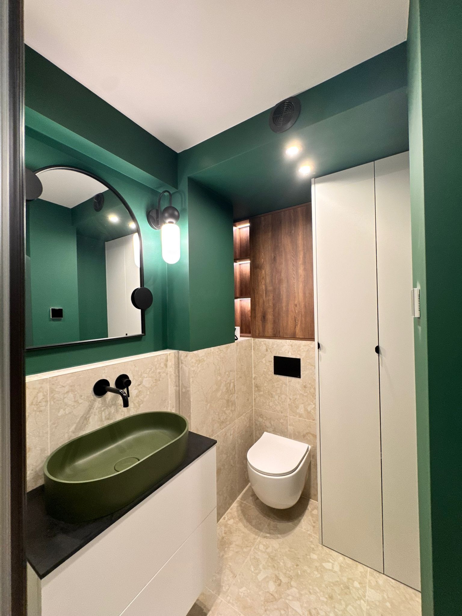 Nowoczesna łazienka z zielonymi ścianami, beżowymi płytkami i umywalką w kolorze oliwkowym. Czarna armatura i lustro z zaokrąglonymi krawędziami dodają elegancji.