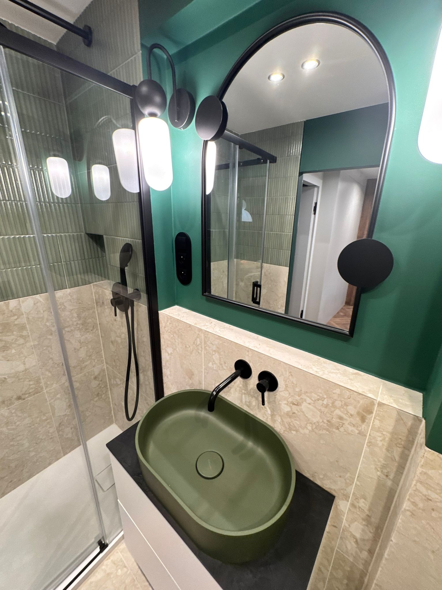 Nowoczesna łazienka z zieloną umywalką nablatową, czarną armaturą i kabiną prysznicową. Ściany w odcieniach zieleni i beżu. Lustro w czarnej ramie.
