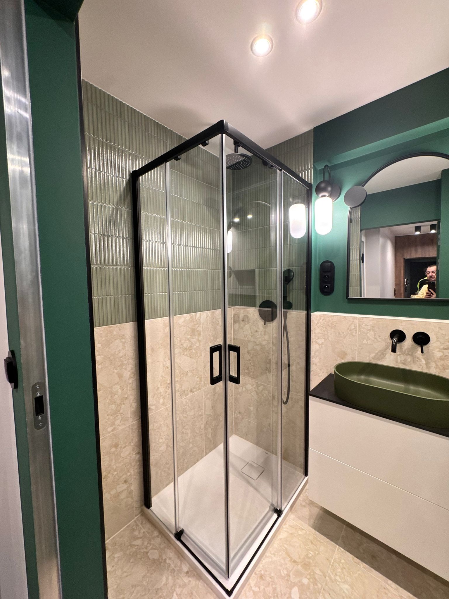 Nowoczesna łazienka z kabiną prysznicową typu walk-in z czarnymi profilami, zieloną umywalką i ścianami w odcieniach zieleni i beżu. Widoczne oświetlenie i lustro.