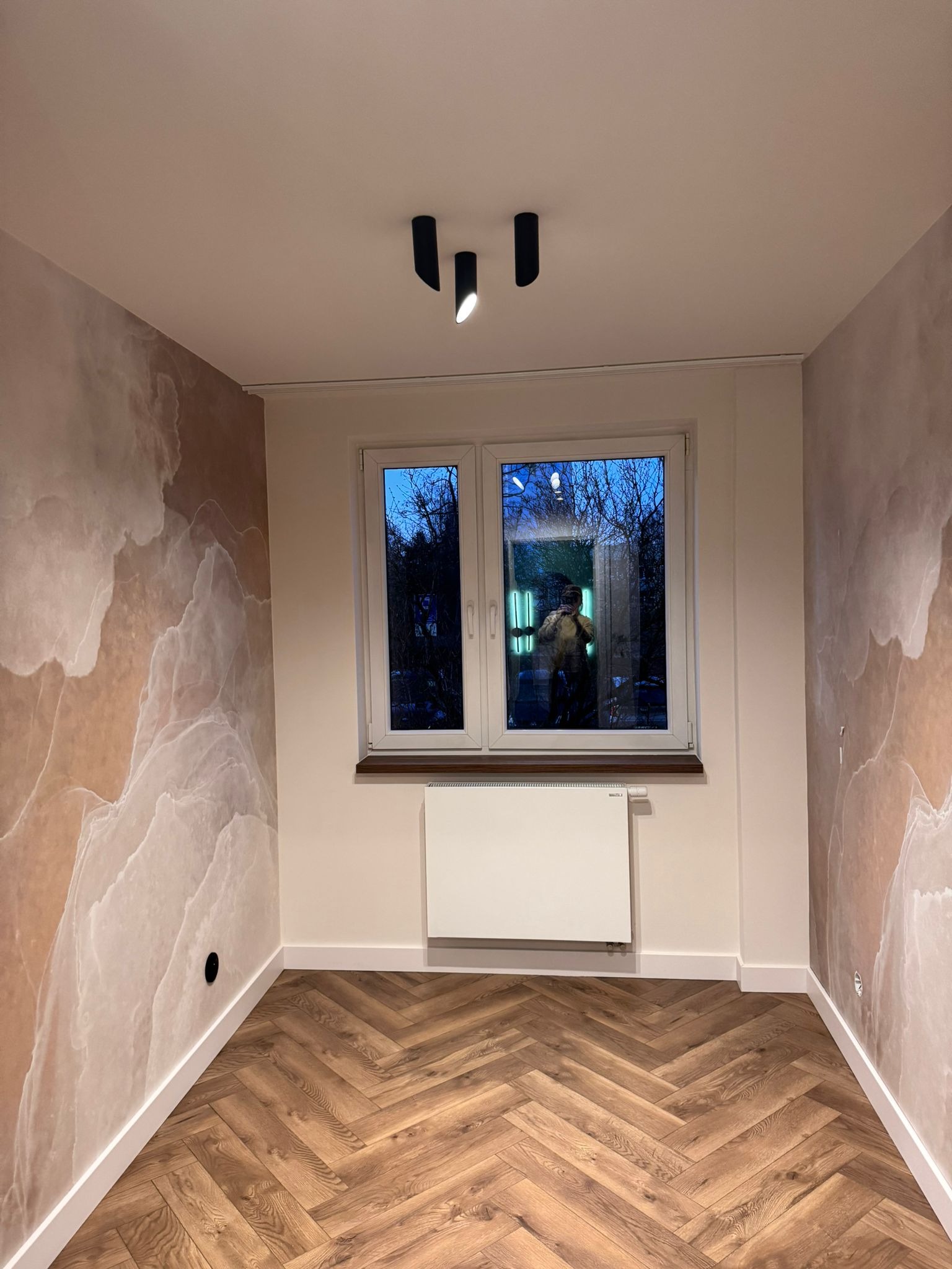 Pokój po remoncie: parkiet w jodełkę, ściany z efektem marmuru, białe okno z widokiem na zewnątrz, nowoczesne oświetlenie sufitowe. Widoczne detale wykończeniowe.