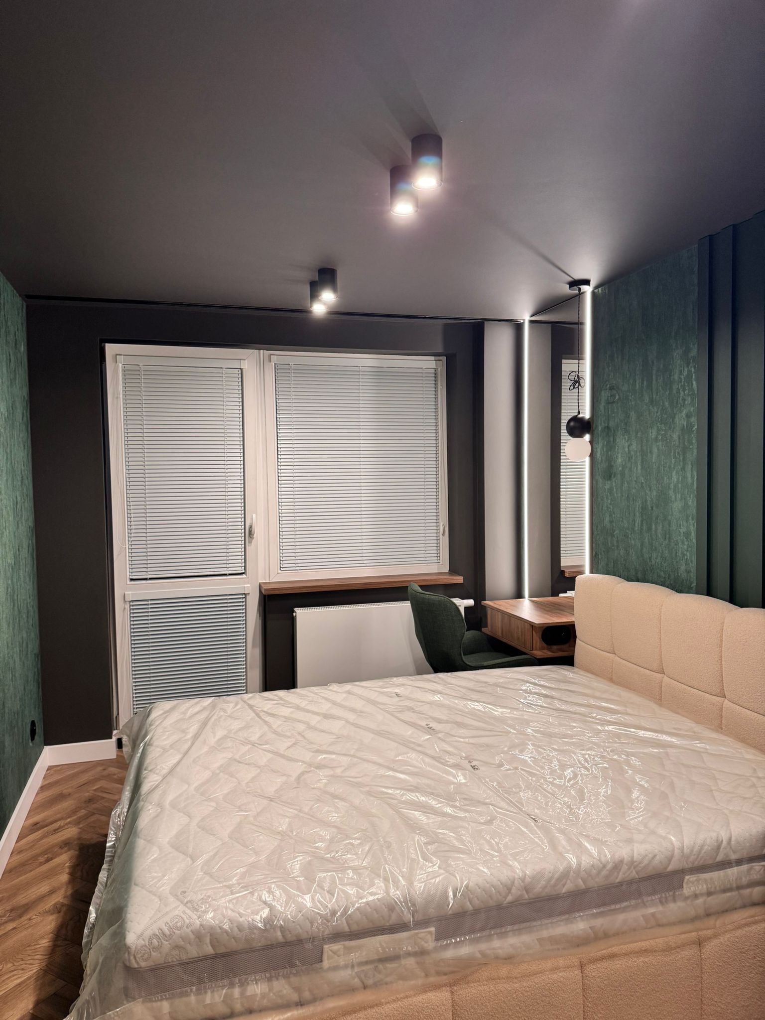 Nowoczesna sypialnia po remoncie: łóżko z beżowym zagłówkiem, biurko z krzesłem, okno z żaluzjami, ciemne ściany z zielonym akcentem, oświetlenie punktowe.
