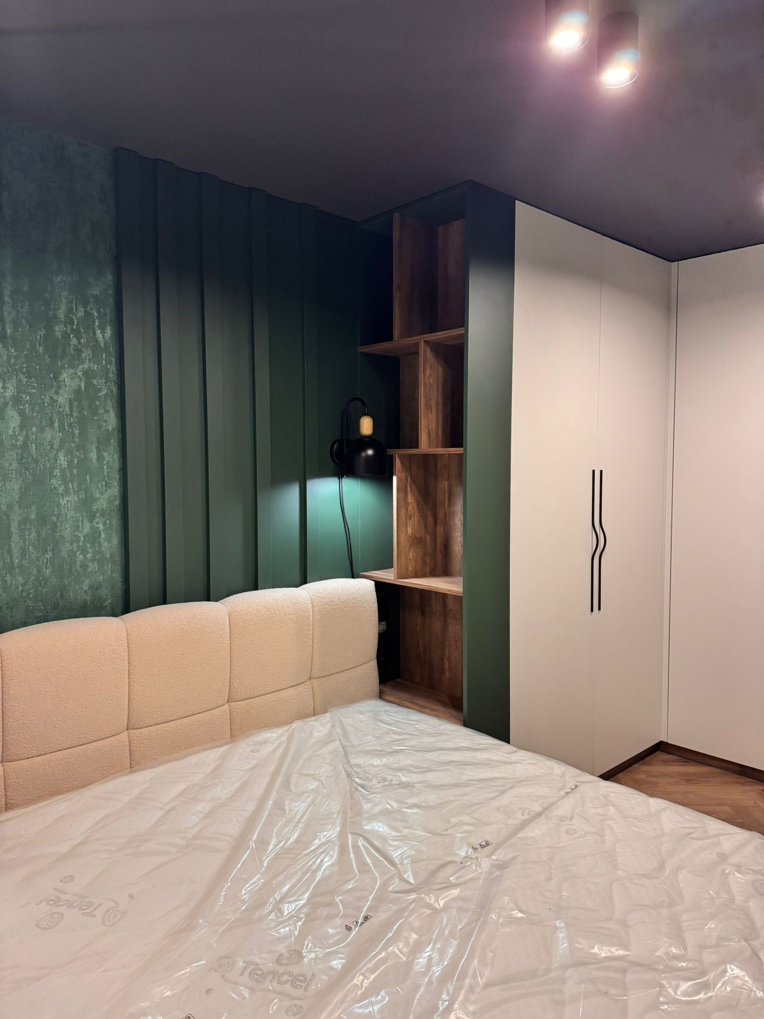 Nowoczesna sypialnia z łóżkiem o beżowym wezgłowiu, zieloną ścianą z lameli, drewnianą półką i białą szafą z czarnymi uchwytami. Całość oświetlona punktowo.