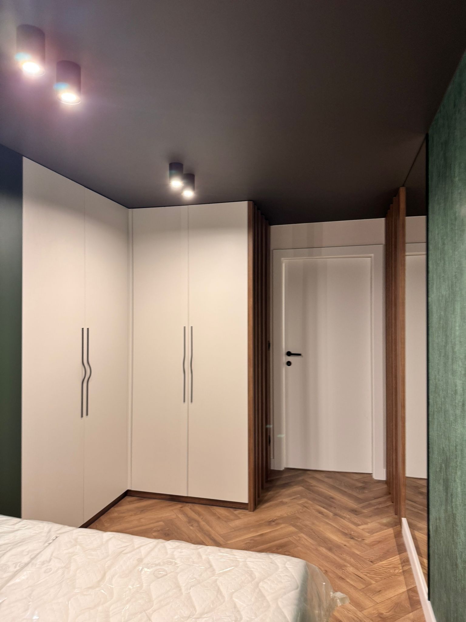 Nowoczesna szafa wnękowa z białymi frontami i minimalistycznymi uchwytami, uzupełniona drewnianymi lamelami. Parkiet w jodełkę i ciemny sufit dodają elegancji.