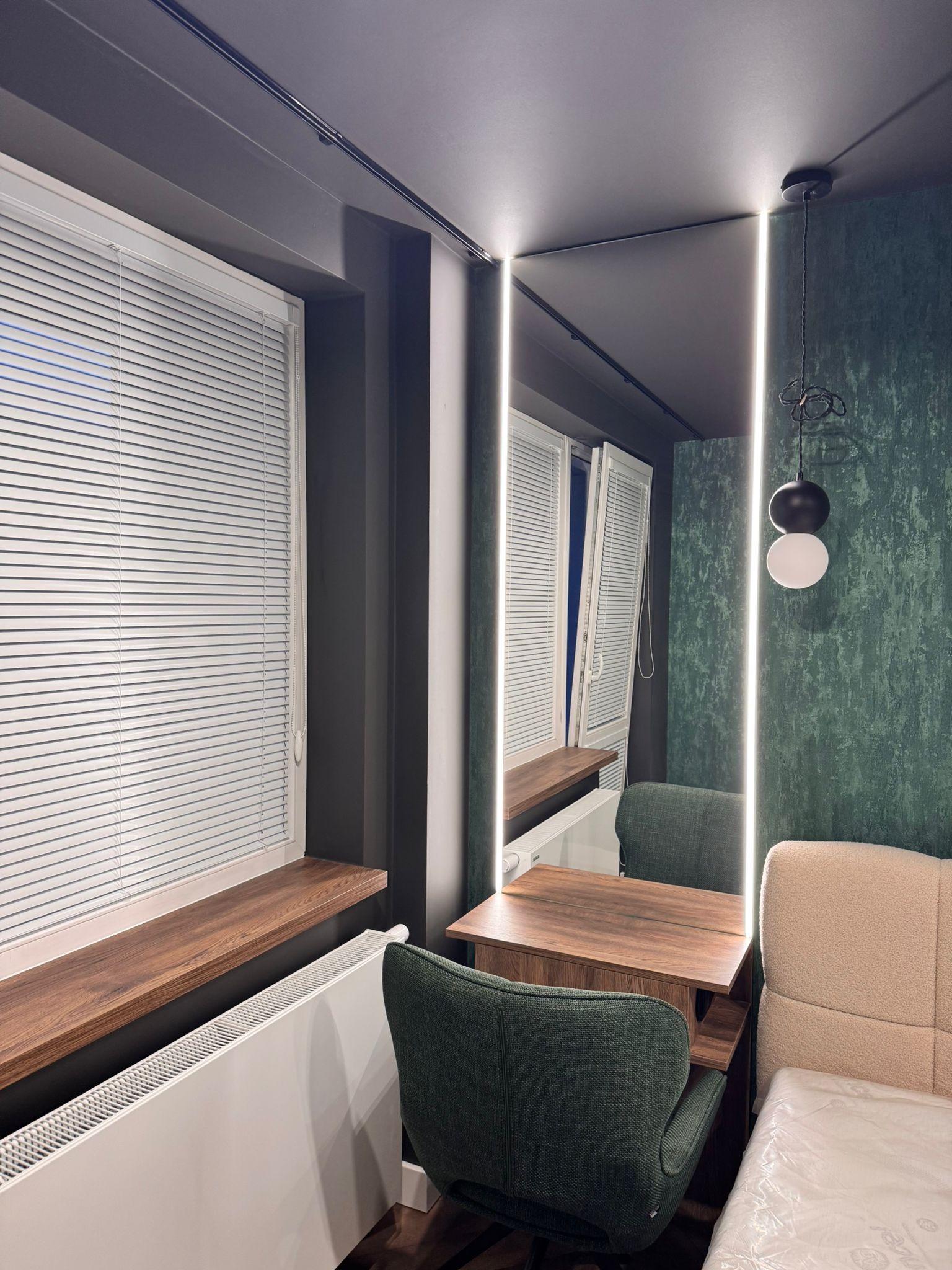 Nowoczesna aranżacja wnętrza: biurko z lustrem z podświetleniem LED, zielone krzesła, okno z żaluzjami i dekoracyjna ściana. Minimalistyczna lampa wisząca.