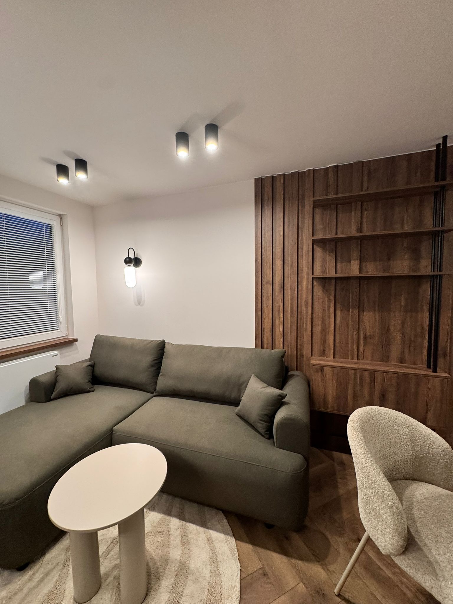 Nowoczesny salon z oliwkową sofą narożną, okrągłym stolikiem i drewnianą zabudową ściany z półkami. Minimalistyczne oświetlenie sufitowe i designerski fotel.