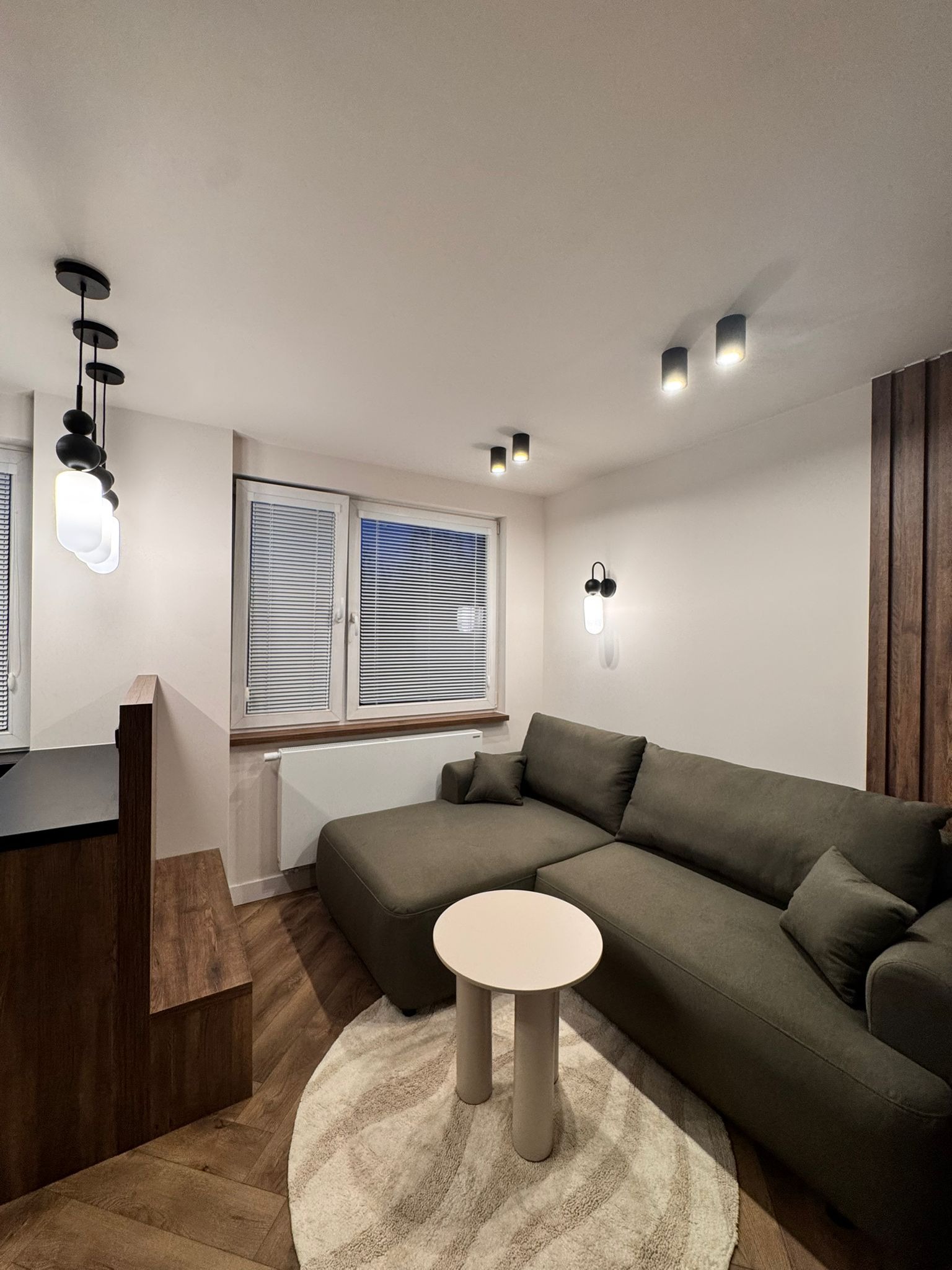 Nowoczesny salon z narożną sofą w odcieniach zieleni, okrągłym stolikiem i drewnianą ścianą. Oświetlenie punktowe i wiszące lampy dodają wnętrzu przytulności.