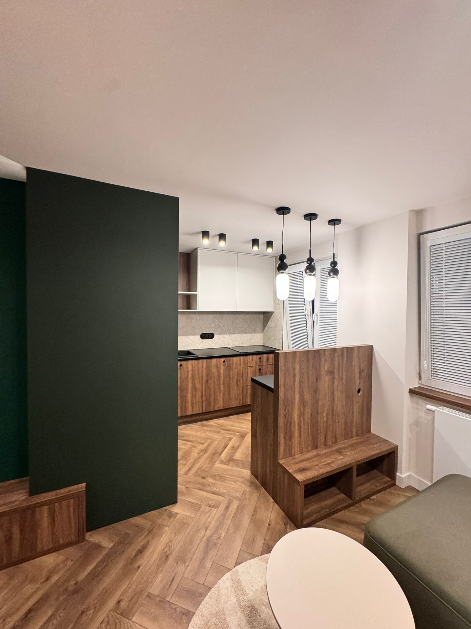 Nowoczesna kuchnia z drewnianymi frontami i białymi szafkami wiszącymi, oddzielona od salonu zieloną ścianką działową i drewnianym barkiem z siedziskiem.