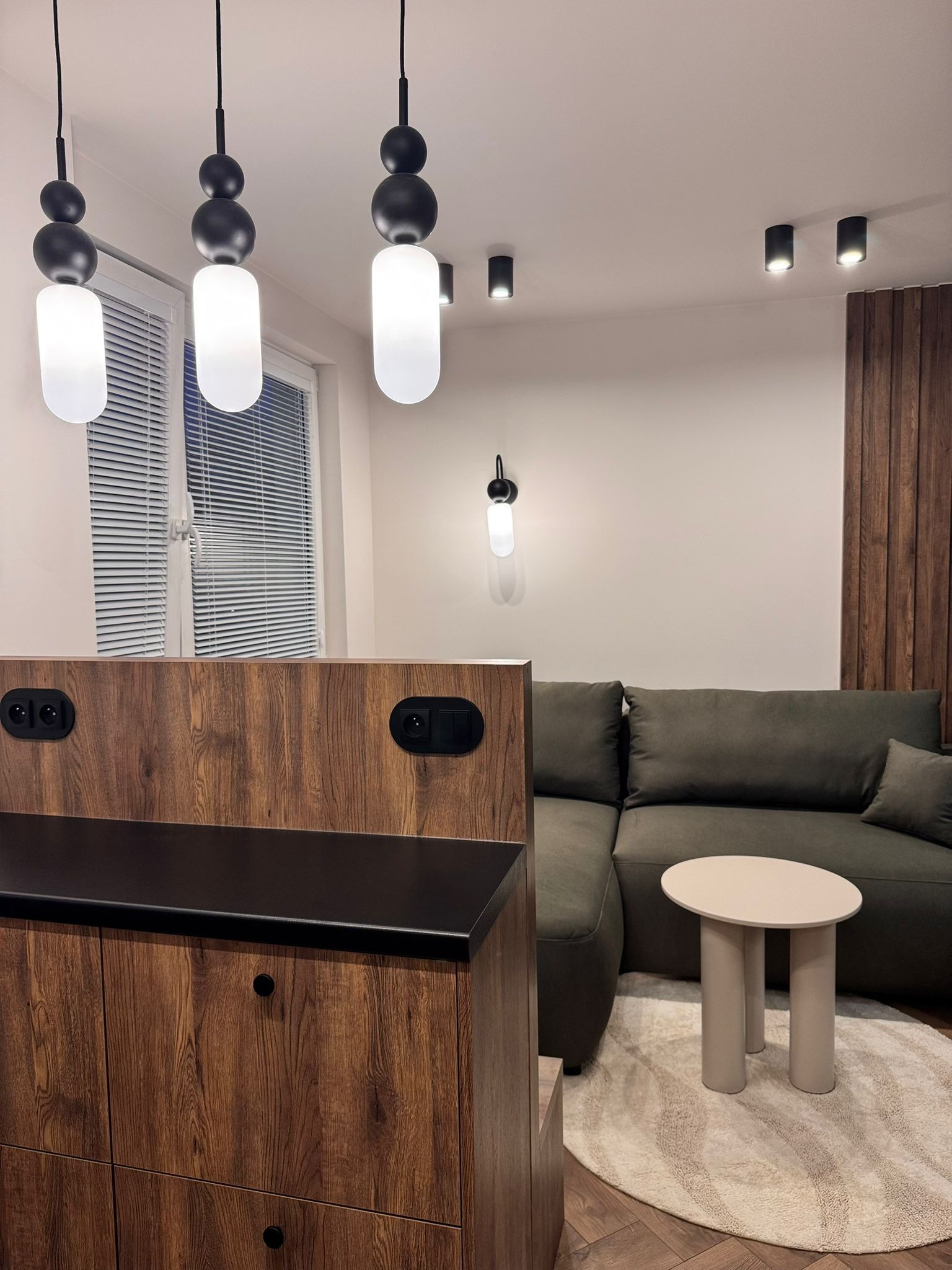 Nowoczesny salon z drewnianą zabudową, szarą sofą i okrągłym stolikiem na wzorzystym dywanie. Oświetlenie stanowią wiszące lampy i reflektory sufitowe.