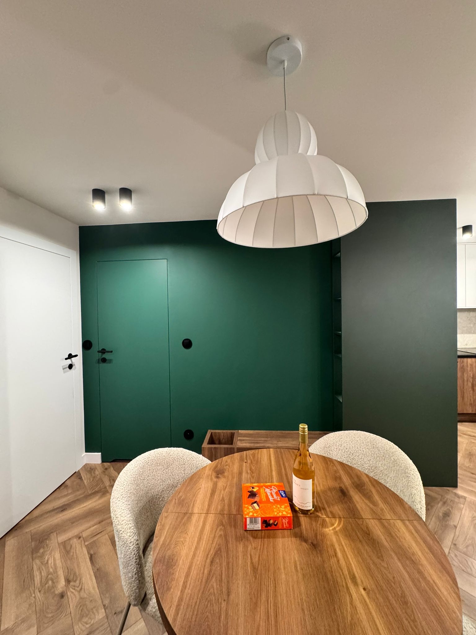 Nowoczesna jadalnia: okrągły drewniany stół z krzesłami boucle, designerska lampa, zielona ściana z ukrytymi drzwiami i parkiet w jodełkę. Minimalistyczny wystrój.