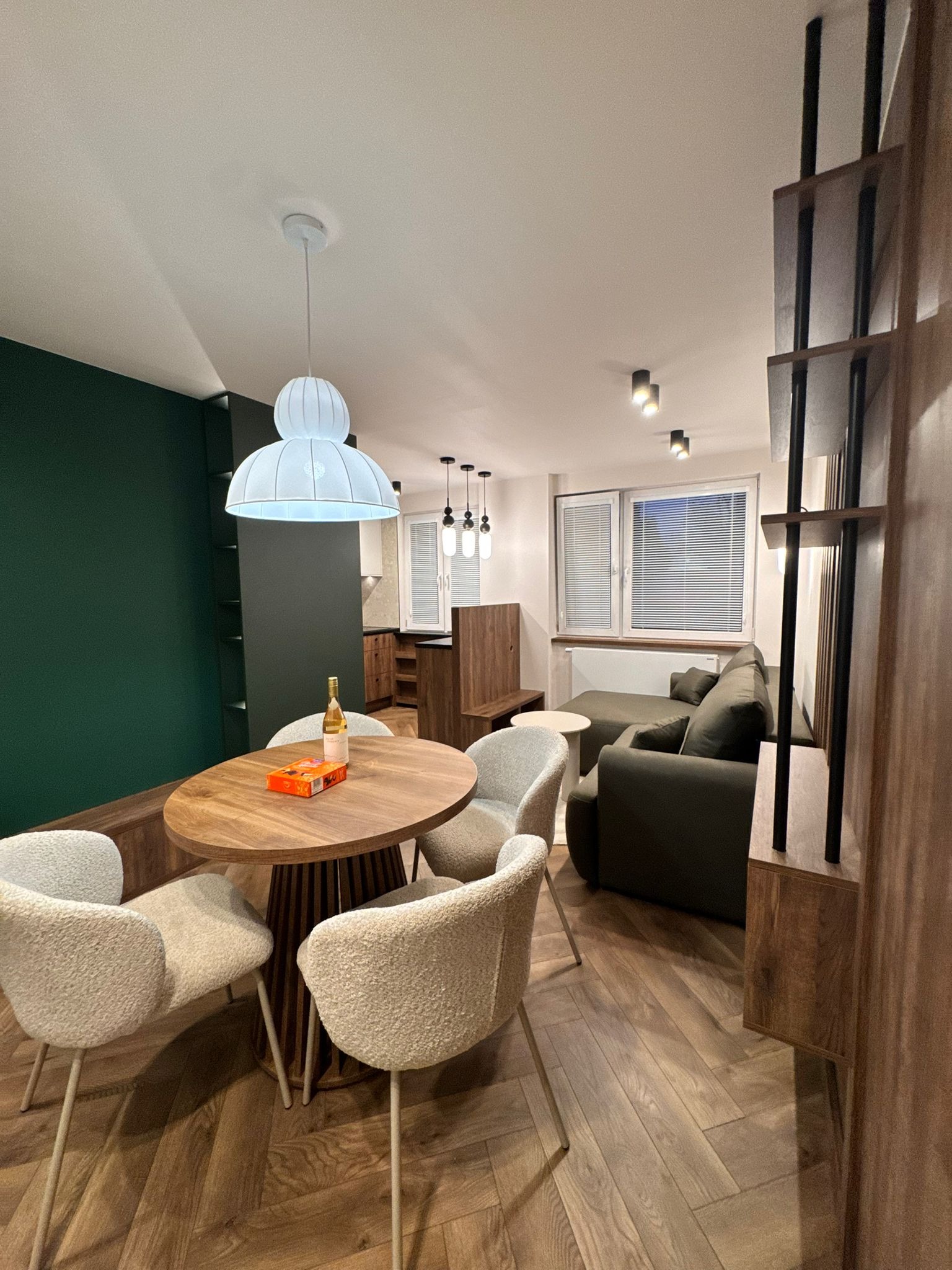 Nowoczesne wnętrze: drewniany stół z krzesłami, zielona ściana, sofa i regał z drewna. Parkiet ułożony w jodełkę. Lampa wisząca nad stołem.