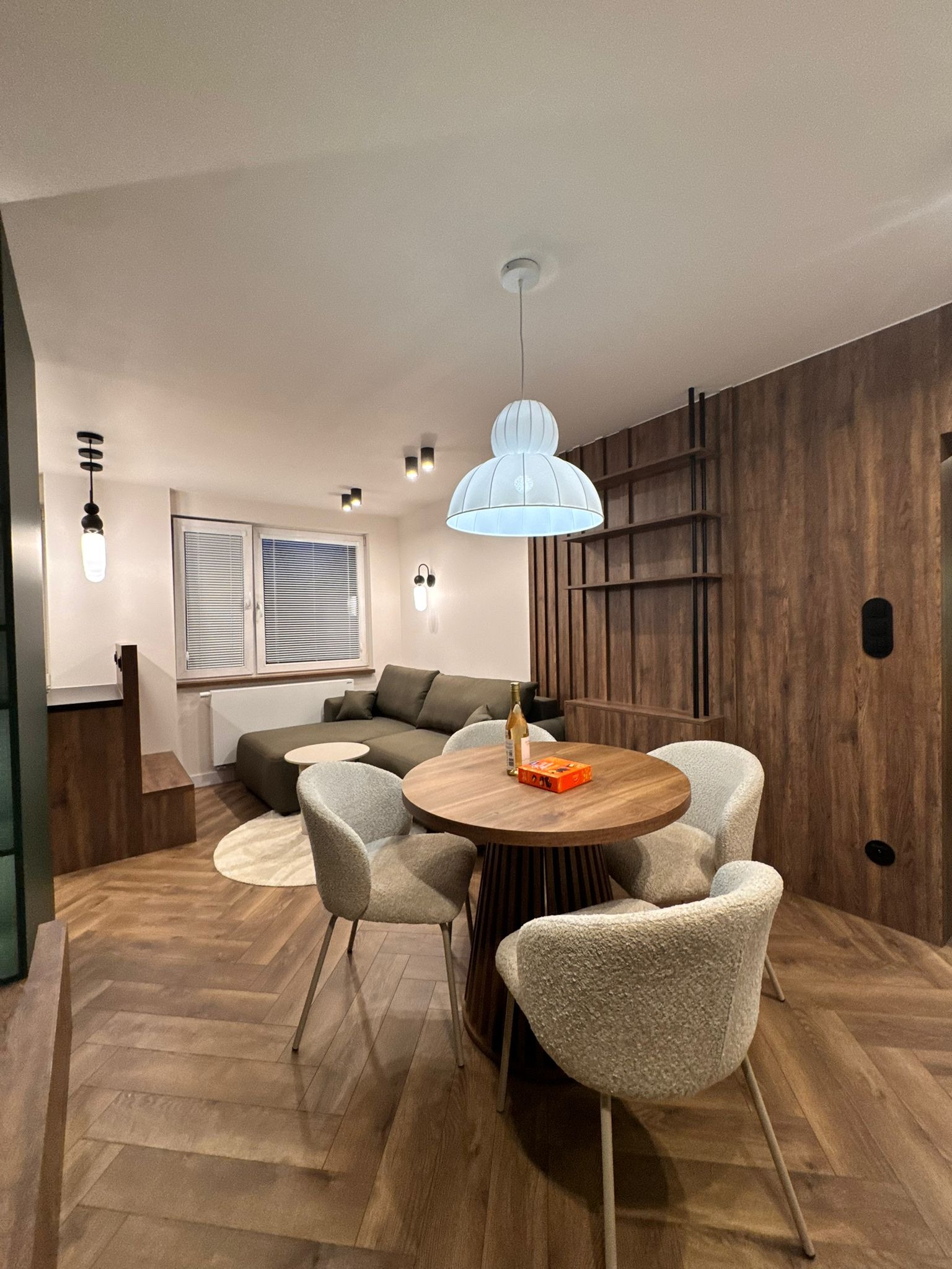 Nowoczesne wnętrze: drewniana podłoga w jodełkę, okrągły stół z krzesłami, sofa w tle, drewniana ściana z półkami i designerskie oświetlenie.