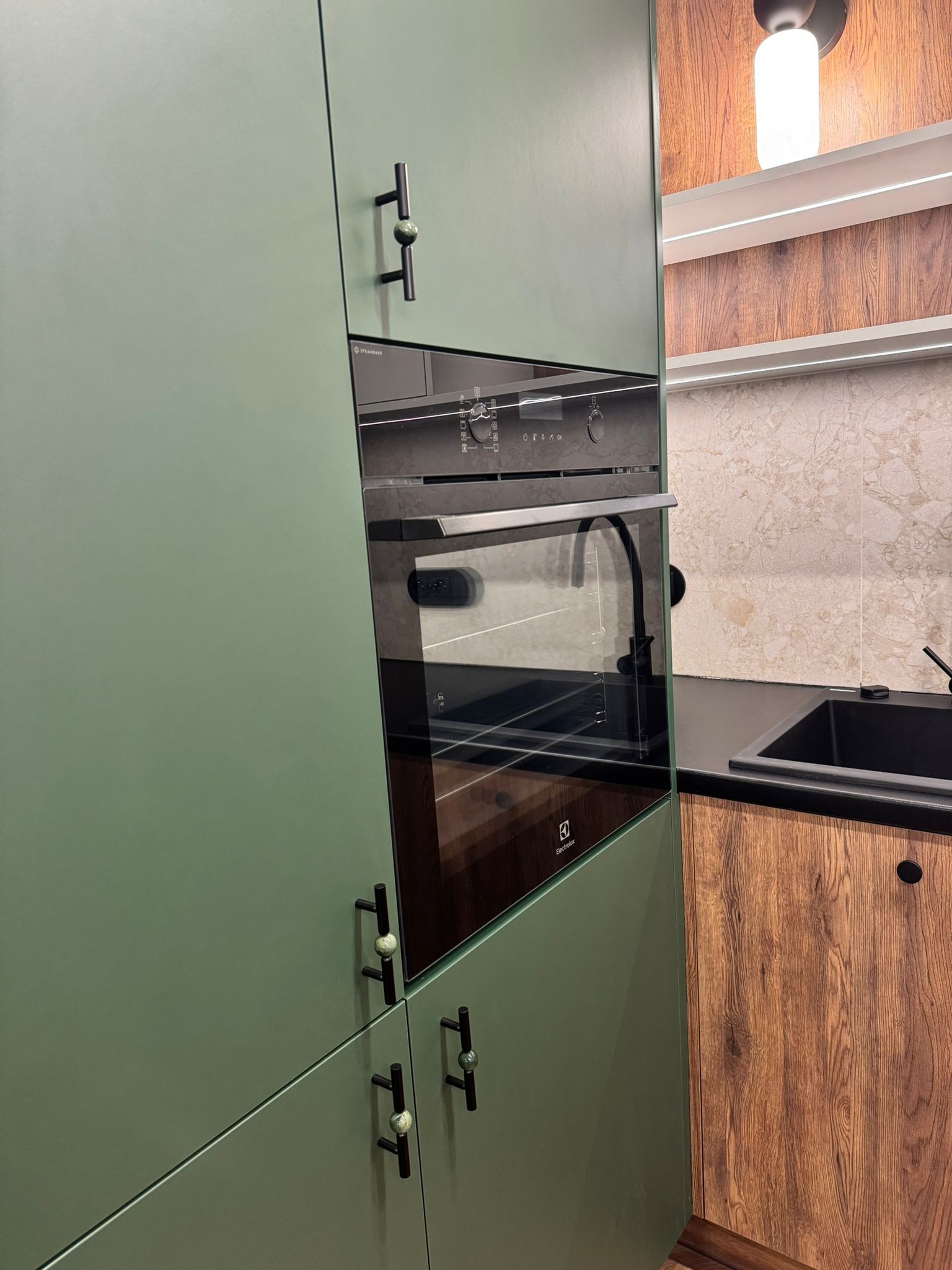 Nowoczesna kuchnia: pistacjowe fronty szafek z czarnymi uchwytami, zabudowany piekarnik Electrolux, drewnopodobne wykończenie, czarny zlew i marmurowe płytki.