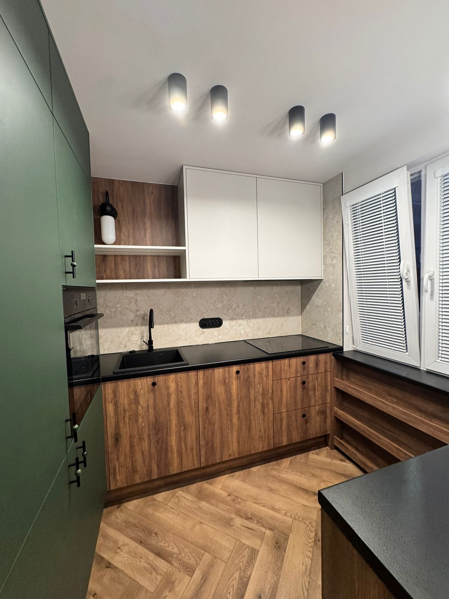 Nowoczesna kuchnia z zielonymi szafkami, drewnianymi frontami i czarnym blatem. Uwagę przykuwa parkiet ułożony w jodełkę i oświetlenie sufitowe.