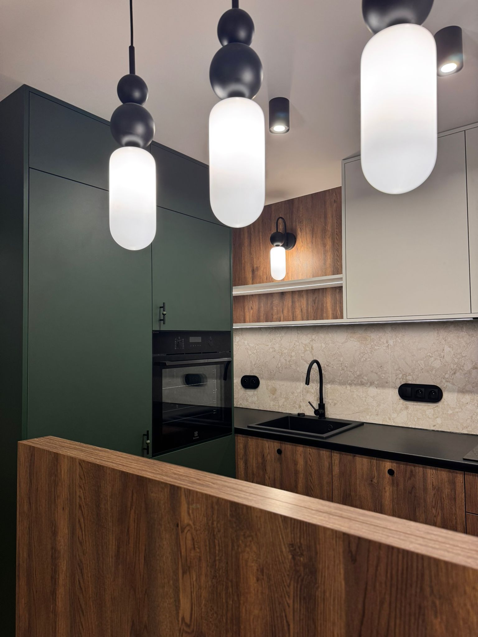 Nowoczesna kuchnia w Gryfowie Śląskim z ciemnozielonymi szafkami, drewnianymi frontami i czarnym blatem. Uwagę przykuwają designerskie lampy wiszące.