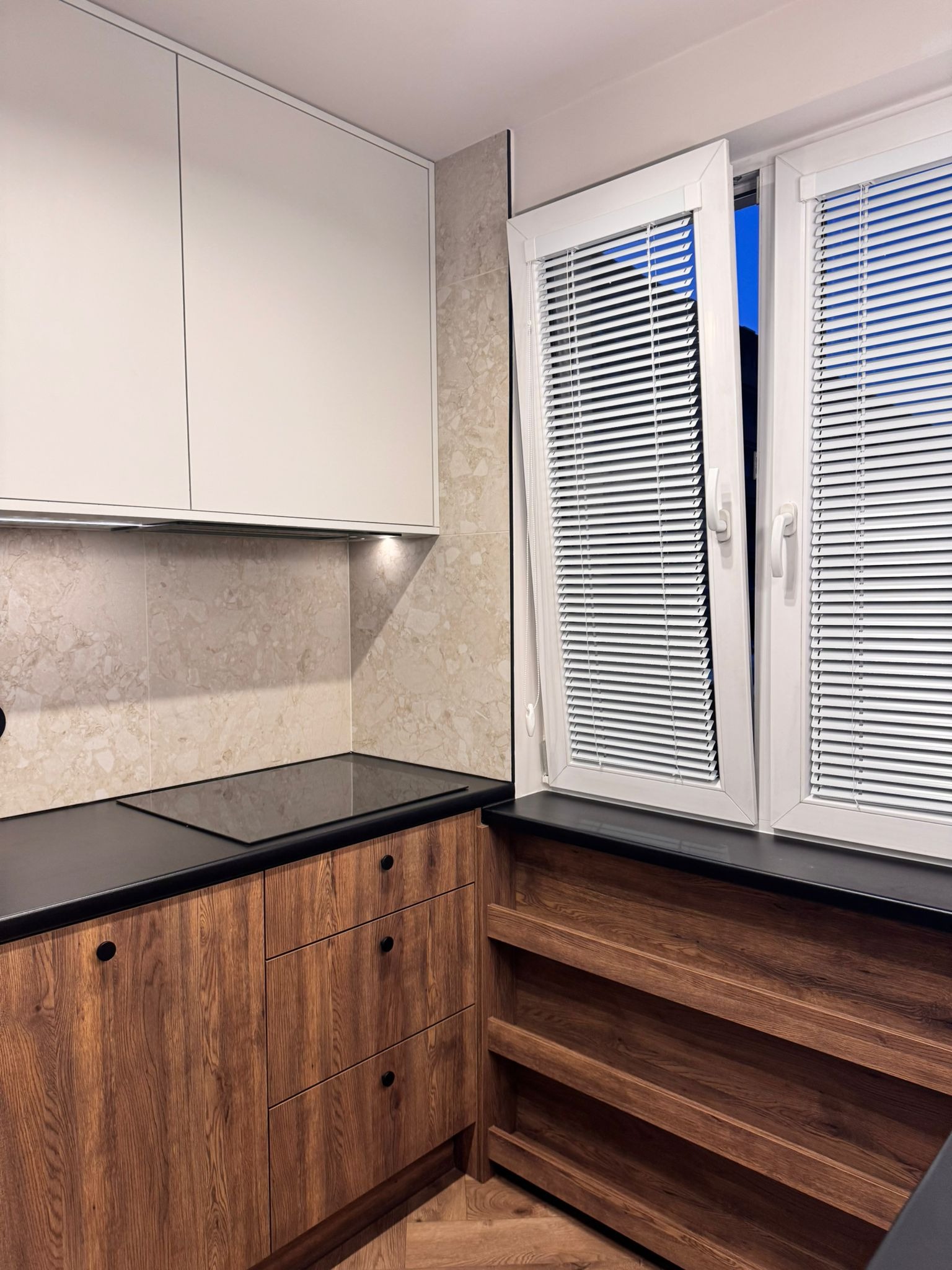 Nowoczesna kuchnia z drewnianymi szafkami, czarnym blatem i białymi górnymi szafkami. Okno z żaluzjami wpuszcza światło. Widoczna płyta indukcyjna i beżowe płytki na ścianie.