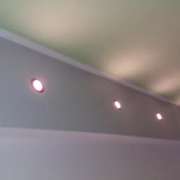 Ściana pomalowana na szaro z zamontowanymi czterema okrągłymi punktowymi lampami LED, oświetlającymi zielony sufit.