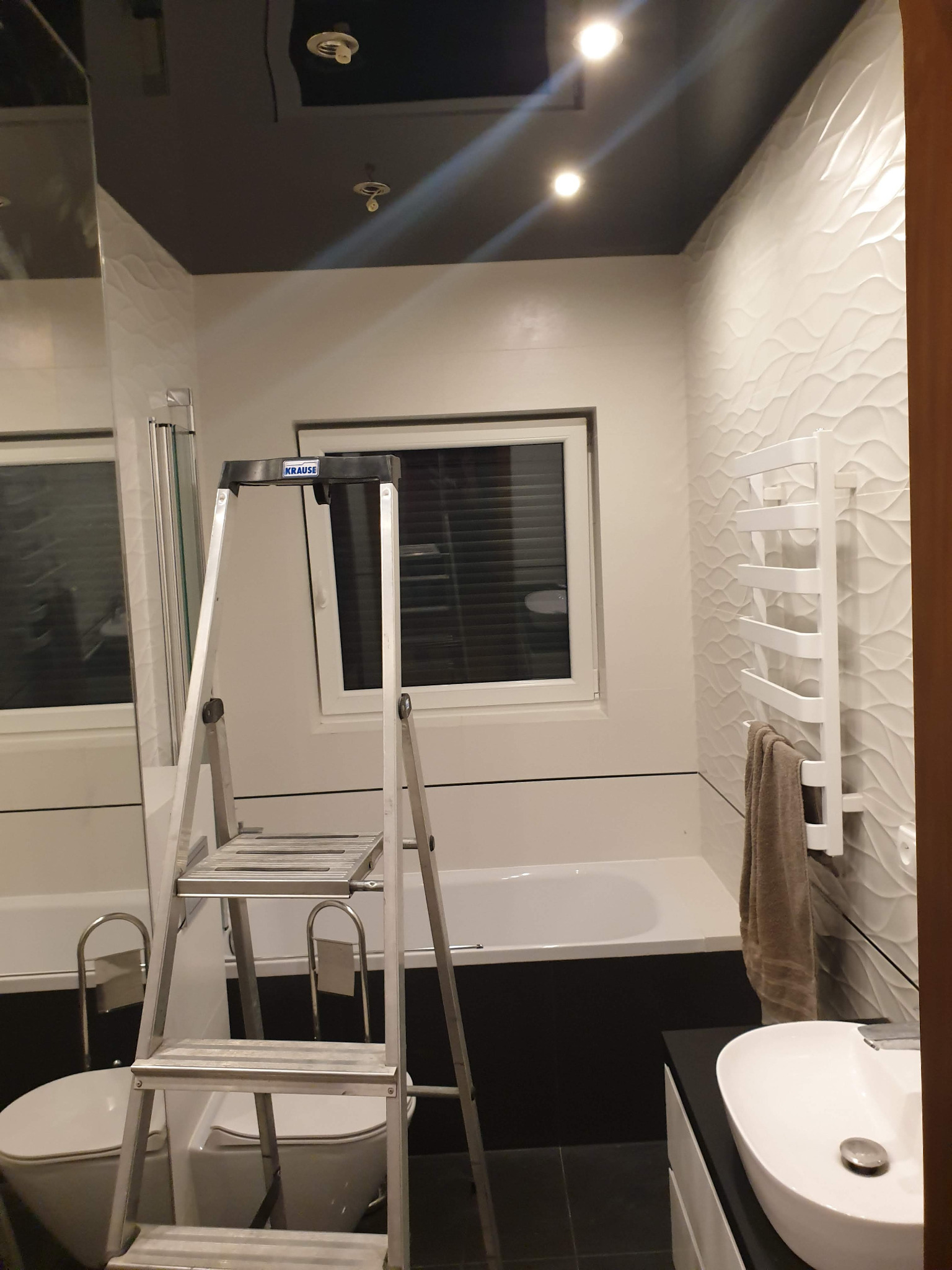 Łazienka w trakcie prac wykończeniowych, widoczna drabina Krause, białe płytki z falistym wzorem, czarny sufit z oświetleniem punktowym, umywalka i grzejnik drabinkowy z ręcznikiem.