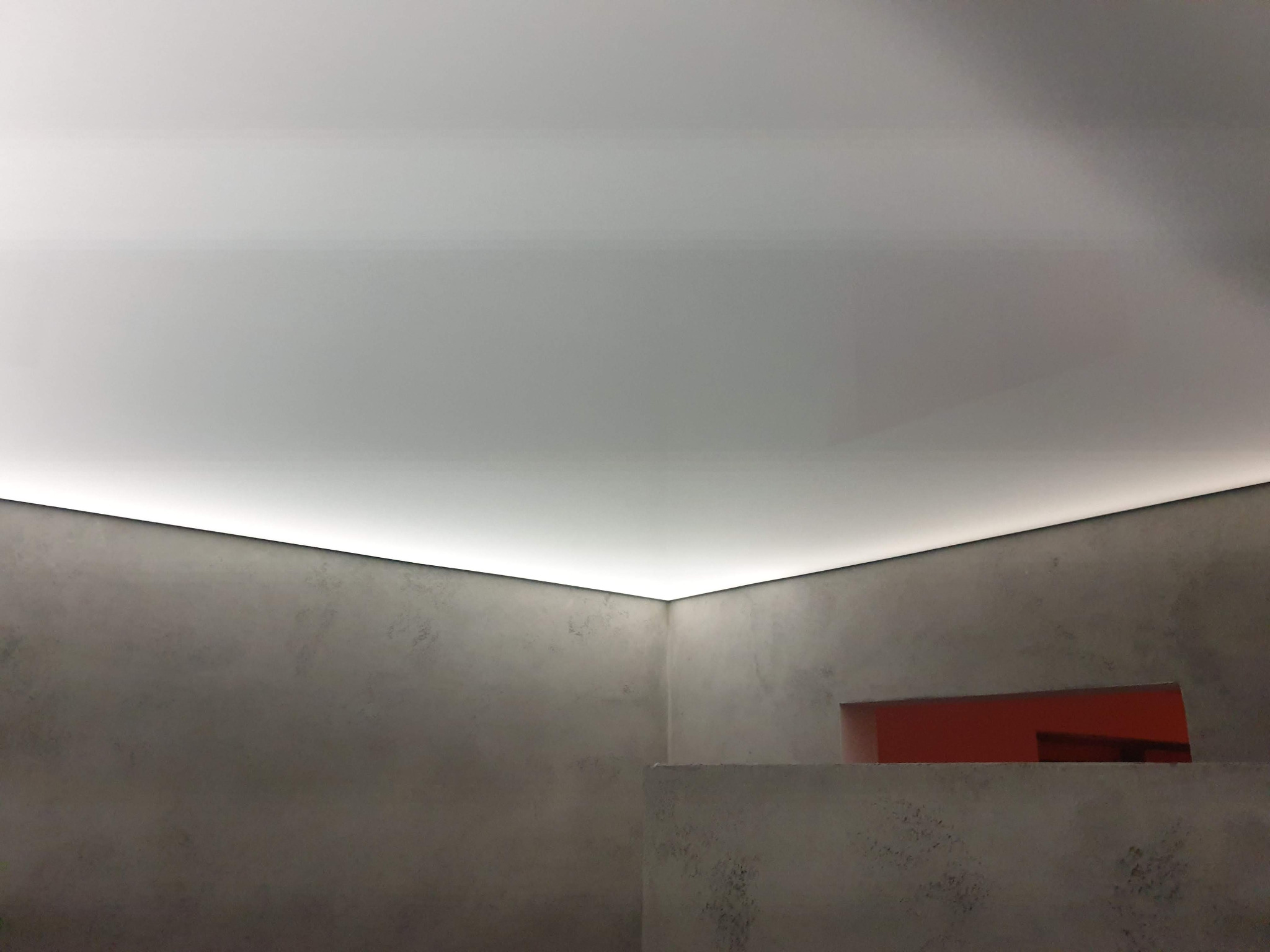 Nowoczesne wnętrze z surowymi szarymi ścianami i podświetlanym sufitem napinanym z ukrytym oświetleniem LED wzdłuż krawędzi, widoczny fragment czerwonej ściany przez otwór w murze.