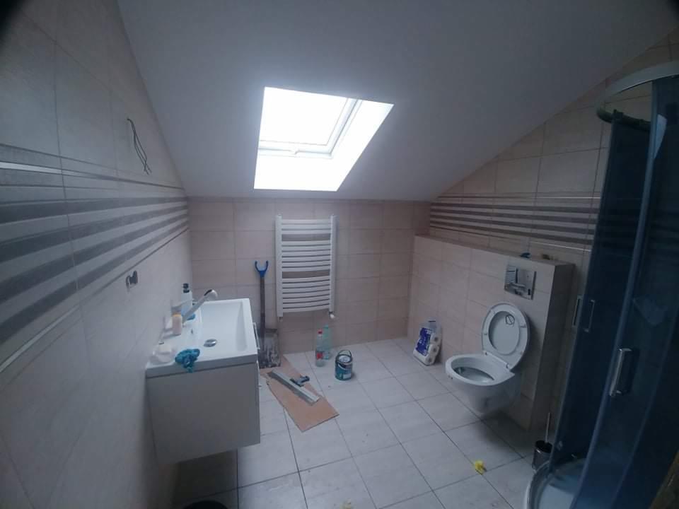 Łazienka na poddaszu w trakcie wykańczania: biała umywalka z szafką, wiszący grzejnik drabinkowy, toaleta, kabina prysznicowa oraz okno dachowe.
