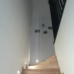 Drewniane schody z podświetleniem LED w ścianie, prowadzące do góry, z czarną balustradą i zdjęciami na ścianie w tle.