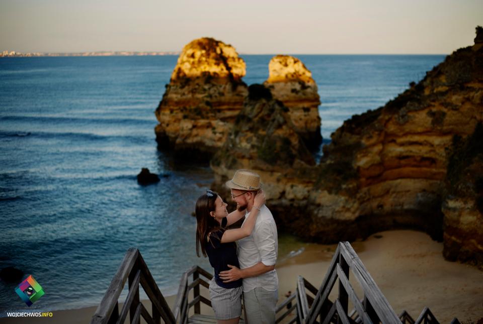 Para obejmująca się na drewnianych schodach z widokiem na morze i formacje skalne w ciepłym świetle zachodzącego słońca.