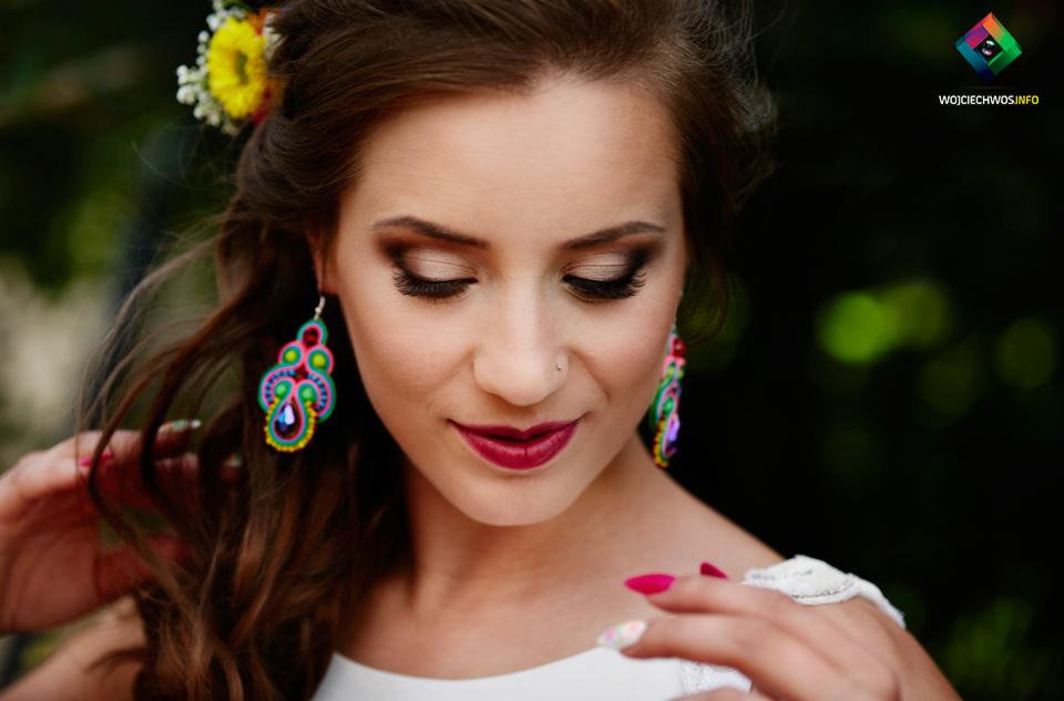 Portret kobiety z kwiatami we włosach i kolorowymi kolczykami, z delikatnym makijażem i manicure, spoglądającej w dół.