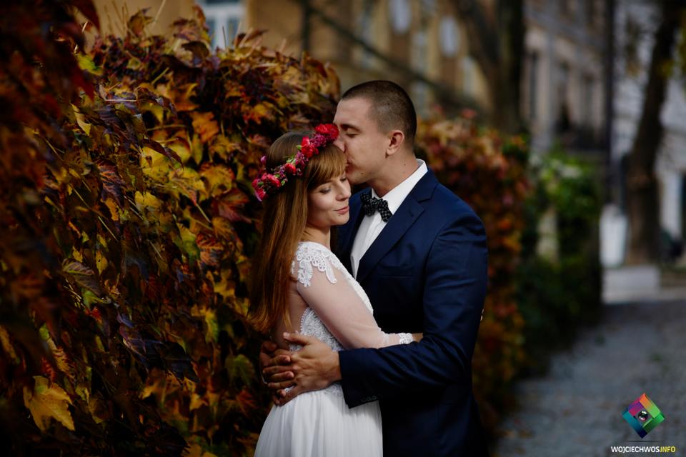 Pan młody całuje pannę młodą w wianku z czerwonych kwiatów na tle jesiennych liści.