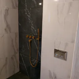 Nowoczesna kabina prysznicowa z czarnymi i białymi płytkami imitującymi marmur, złota armatura, wisząca miska WC z chromowanym przyciskiem spłukującym.