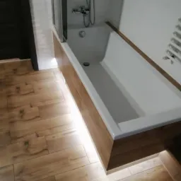 Nowoczesna obudowa wanny z drewnopodobnych paneli z podświetleniem LED, uzupełniająca aranżację łazienki z płytkami imitującymi drewno na podłodze i białą armaturą.