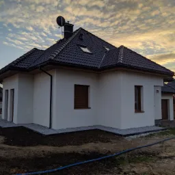 Nowoczesny, parterowy dom z ciemną dachówką i białymi ścianami, widok z zewnątrz po zakończeniu prac, z widocznym fragmentem ogrodu w trakcie zagospodarowywania.