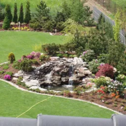 Widok z góry na zadbany ogród z wodospadem kaskadowym z kamieni, otoczonym kwitnącymi krzewami i drzewami iglastymi, graniczący z równo przystrzyżonym trawnikiem i ogrodzeniem.