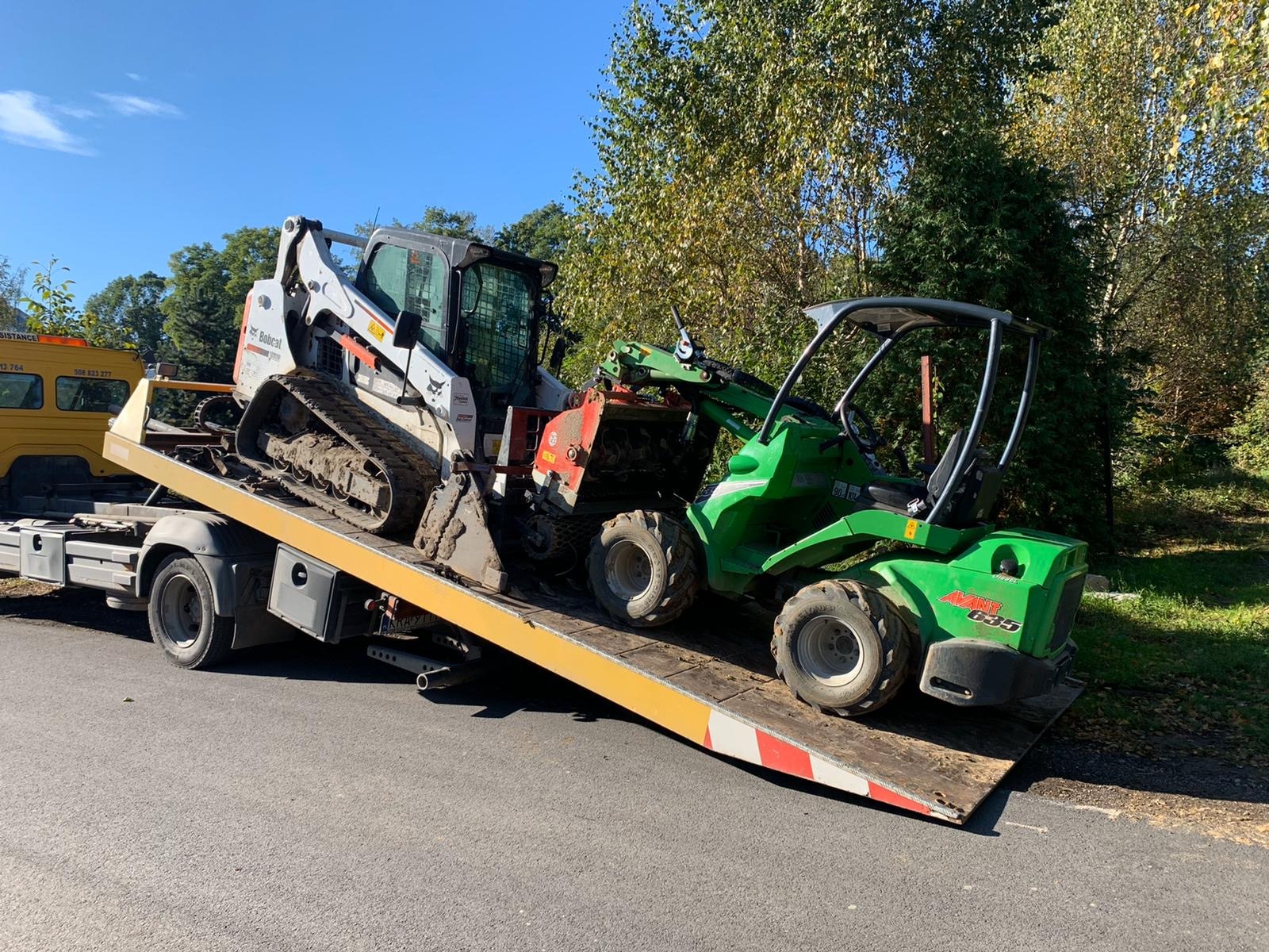 Transport dwóch maszyn budowlanych: białej miniładowarki gąsienicowej Bobcat i zielonej ładowarki przegubowej Avant 635, na żółtej lawecie holowniczej, na tle drzew i błękitnego nieba.