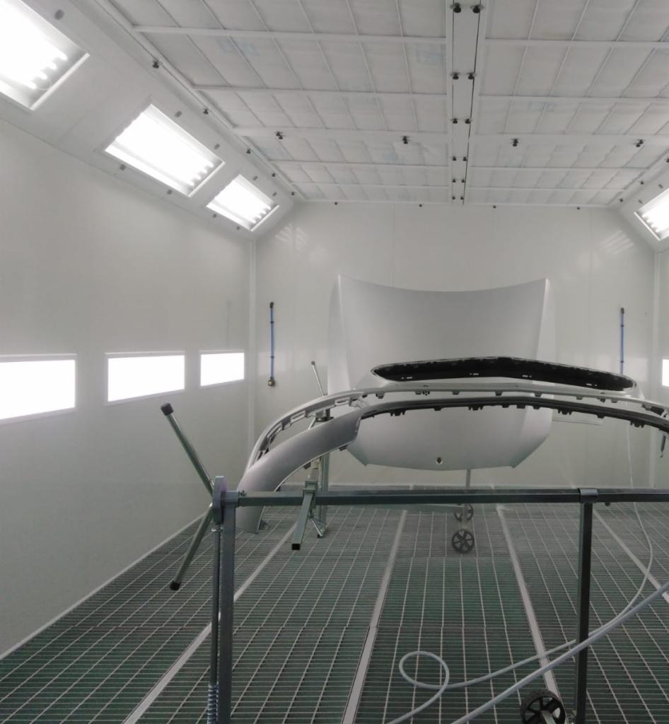 Elementy karoserii samochodowej, w tym zderzak i maska, przygotowane do lakierowania w profesjonalnej kabinie lakierniczej z kratą podłogową i oświetleniem sufitowym.