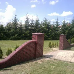 Ceglana brama wjazdowa z niskim murem, wykończona metalowymi słupkami. W tle rząd iglaków i błękitne niebo z chmurami. Widoczny fragment chodnika i sterta piasku.