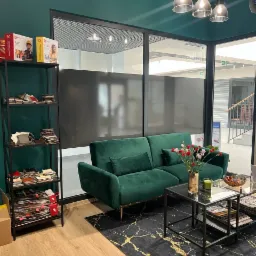 Szmaragdowa sofa dwuosobowa z aksamitną tapicerką, ustawiona na czarnym dywanie ze złotymi żyłkami, obok regał z próbkami materiałów tapicerskich, w tle przeszklona ściana biurowa.