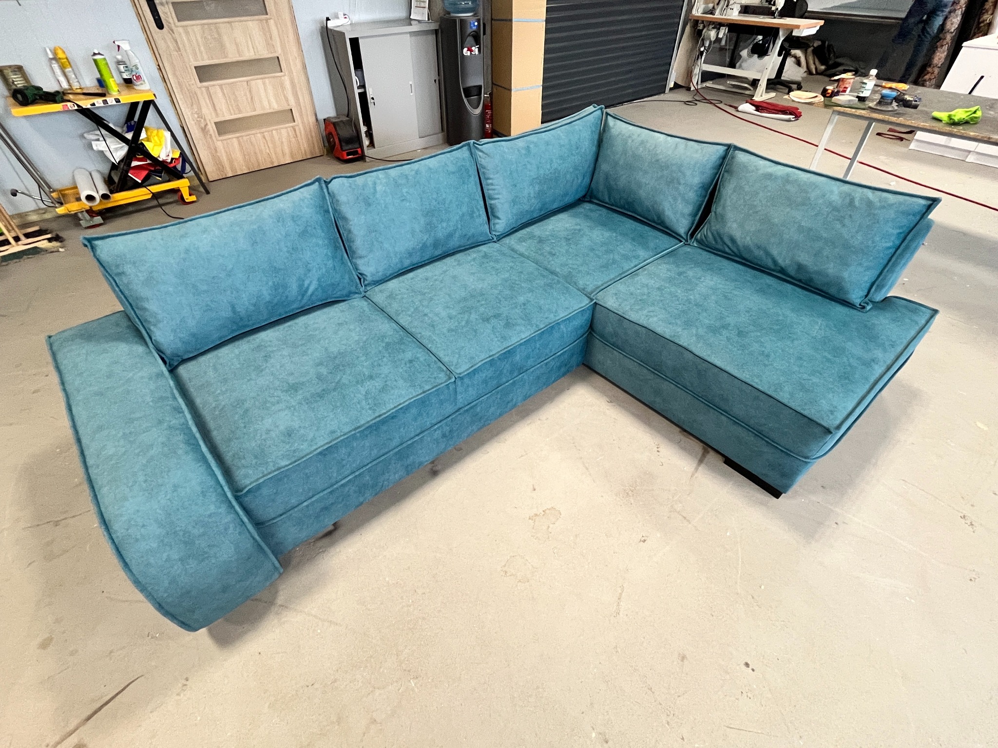 Narożnik tapicerowany w odcieniach turkusu, z widocznymi szwami i przeszyciami, ustawiony w warsztacie z widocznym wyposażeniem i narzędziami.