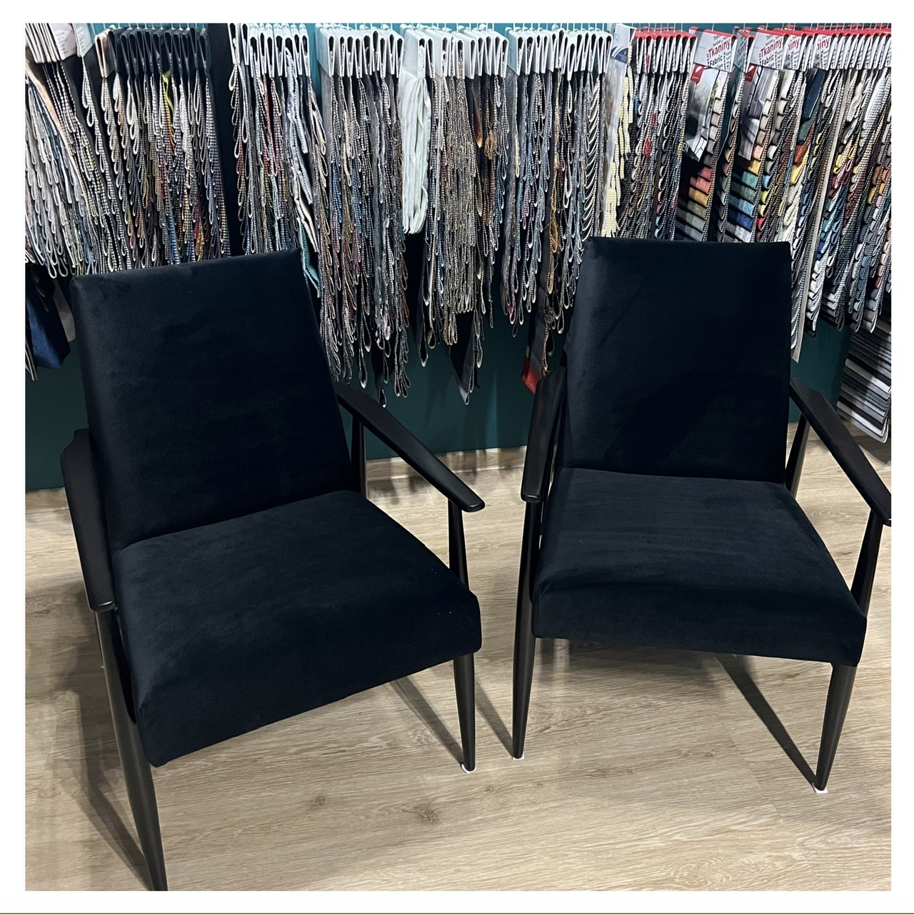 Dwa identyczne, czarne, welurowe fotele z drewnianymi podłokietnikami i nogami na tle ściany z wiszącymi próbkami tkanin.