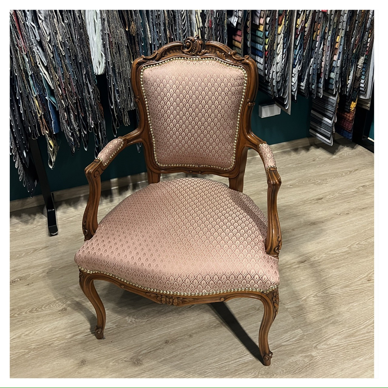Odrestaurowany fotel w stylu Ludwika XVI z różową tapicerką w drobny wzór, na tle ekspozycji tkanin obiciowych