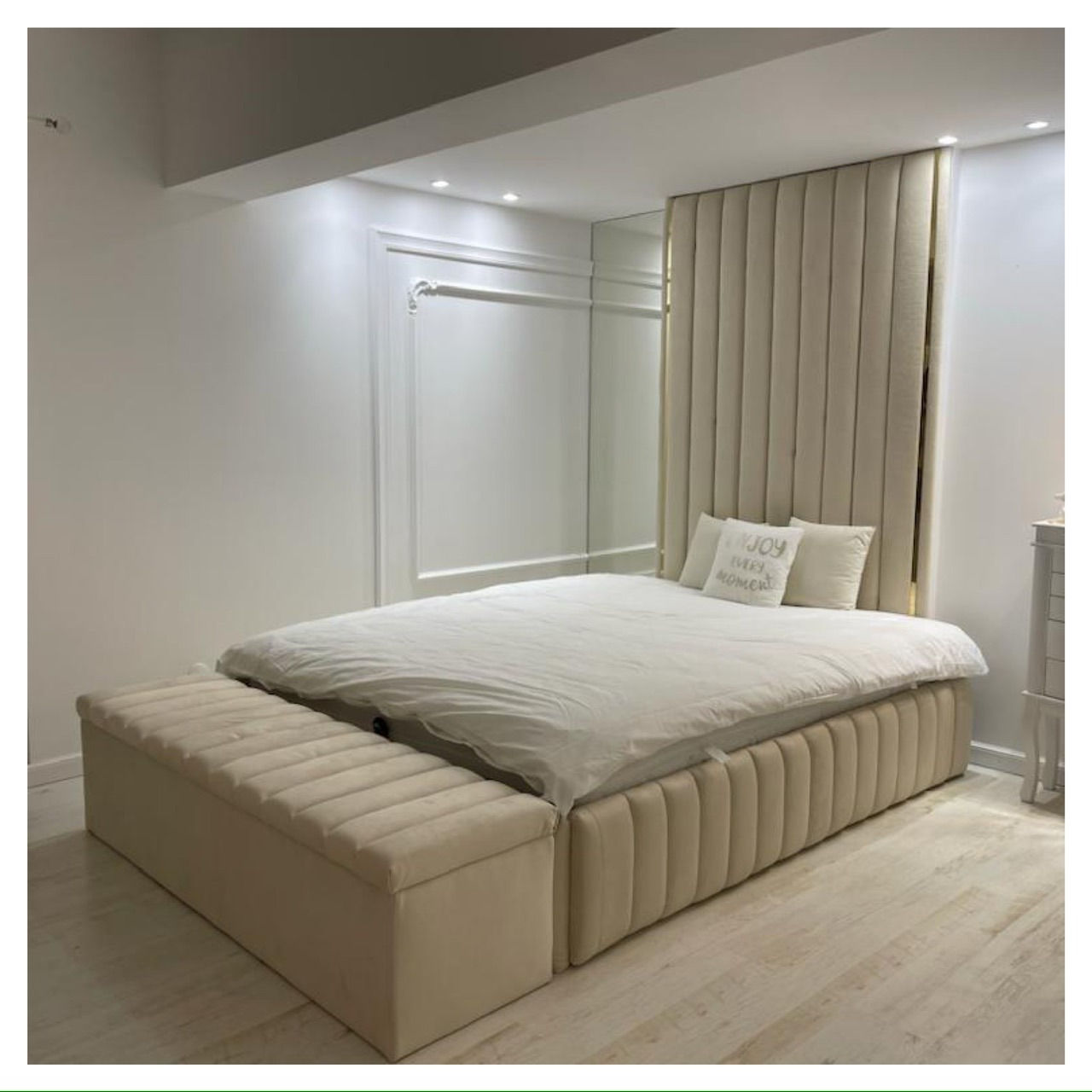 Beżowe łóżko tapicerowane z wysokim zagłówkiem z pionowymi przeszyciami i skrzynią, uzupełnione o dwie białe poduszki z napisem, ustawione na drewnianej podłodze w minimalistycznej sypialni...