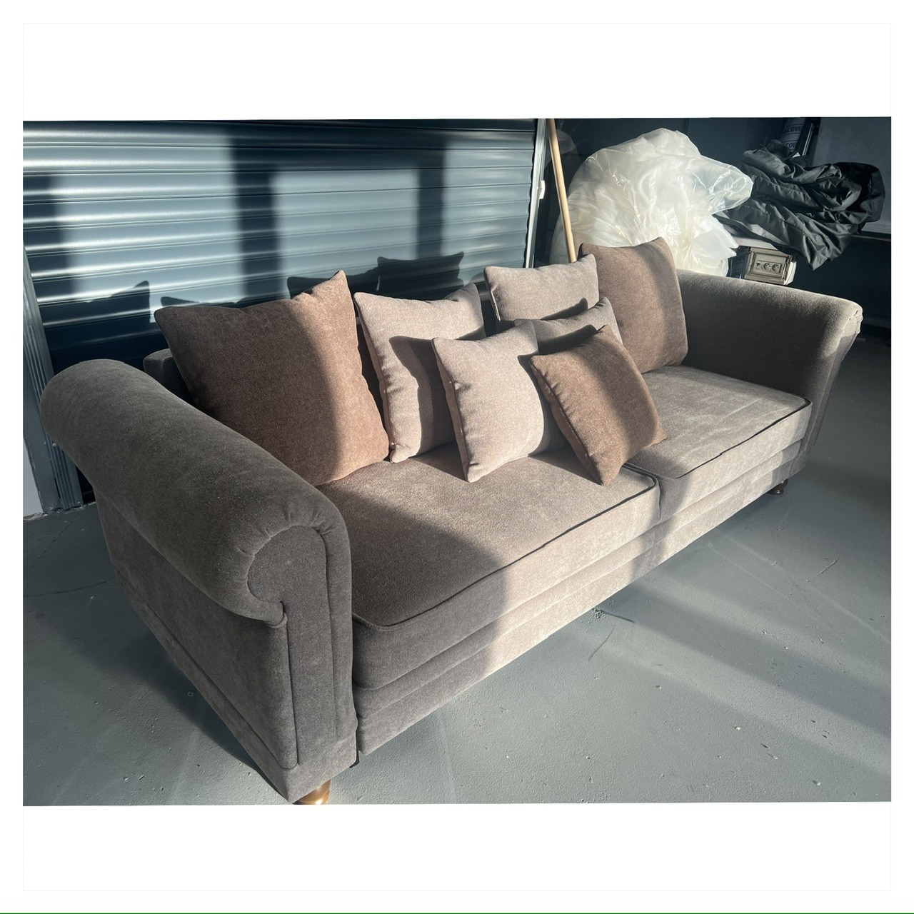 Szara sofa z poduszkami w odcieniach beżu i brązu, ustawiona na szarej podłodze na tle zamkniętej, metalowej rolety garażowej.