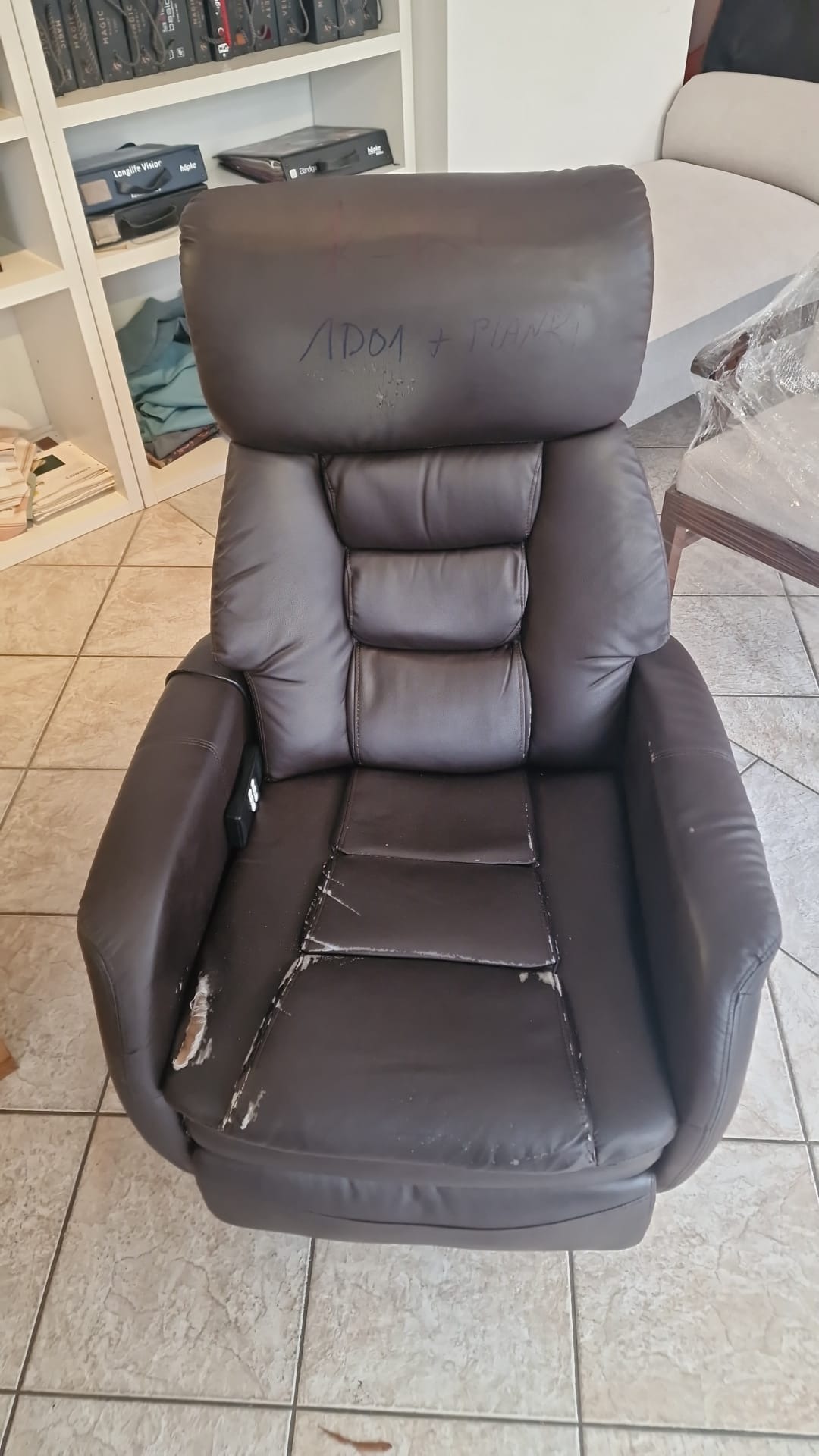 Zniszczony, brązowy fotel tapicerowany z widocznymi przetarciami i napisem 'APDA + PIANKI' na zagłówku, stojący na kafelkach w pomieszczeniu z regałem na książki w tle.