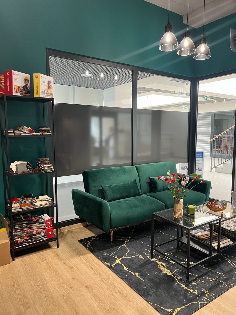 Szmaragdowa sofa dwuosobowa z aksamitną tapicerką, ustawiona na czarnym dywanie ze złotymi żyłkami, obok regał z próbkami materiałów tapicerskich, w tle przeszklona ściana biurowa.
