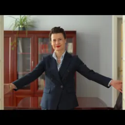 Kobieta w granatowym garniturze otwiera białe, przeszklone drzwi, za nią widoczny gabinet z drewnianą biblioteczką i biurkiem.