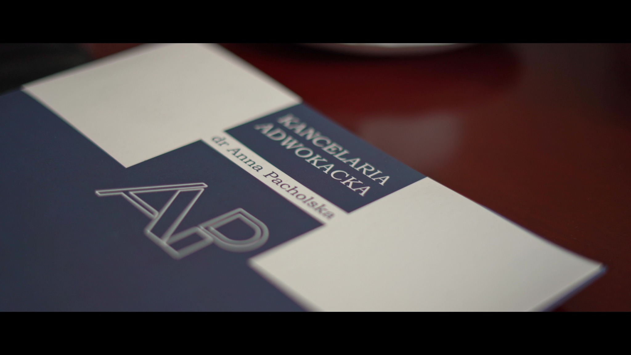 Wizytówka Kancelarii Adwokackiej dr Anny Pacholskiej z minimalistycznym logo AP na granatowym tle, leżąca na ciemnym blacie.