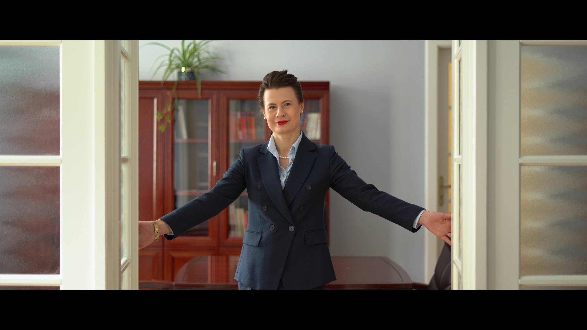 Kobieta w granatowym garniturze otwiera białe, przeszklone drzwi, za nią widoczny gabinet z drewnianą biblioteczką i biurkiem.