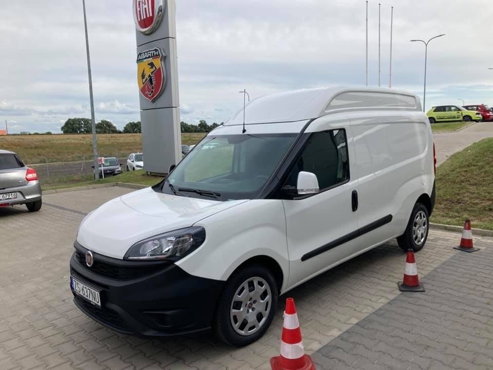 Biały samochód dostawczy Fiat Doblo z podwyższonym dachem na parkingu, widoczny znak dealera Fiat i Abarth w tle, otoczony pachołkami ostrzegawczymi.