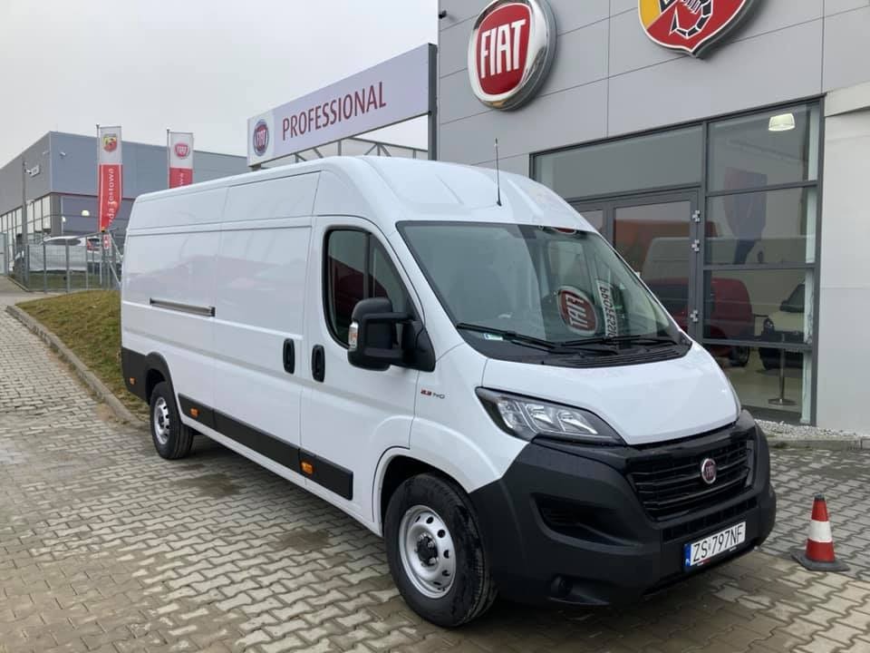Biały furgon Fiat Professional zaparkowany przed salonem samochodowym, widoczny znak dealera i pachołek ostrzegawczy.