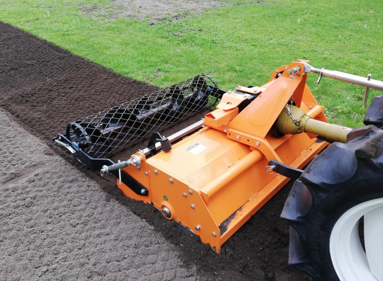 Pomarańczowa glebogryzarka z wałem strunowym wyrównuje świeżo przekopaną ziemię, przygotowując teren pod siew trawy. Widoczny fragment ciągnika.