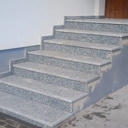 Usługi Kamieniarskie Kamil Kopras - Zewnętrzne schody z szarego kamienia prowadzące do drzwi wejściowych, z widocznymi śladami poprawek tynkarskich na ścianie i podłodze.