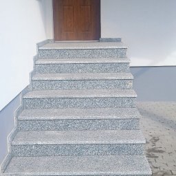 Usługi Kamieniarskie Kamil Kopras - Zewnętrzne schody z szarego kamienia prowadzące do drewnianych drzwi wejściowych, z widoczną kostką brukową na podeście.